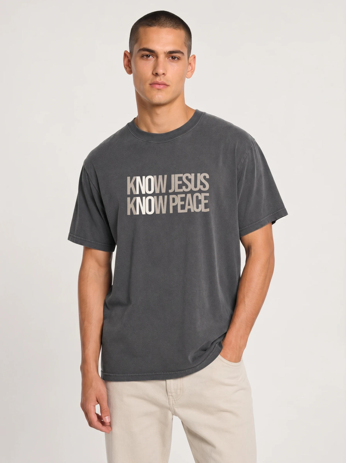 A man wearing a dark gray washed graphic t-shirt with the text 'KNOW JESUS KNOW PEACE' in white, paired with light beige pants, against a light gray studio background.