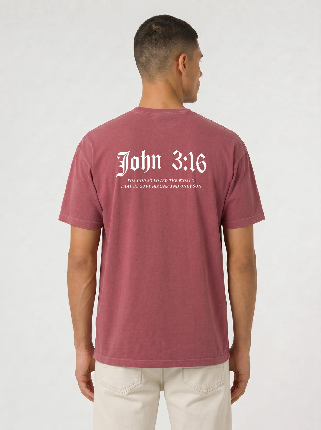 A man viewed from the back wearing a heathered dusty rose t-shirt with 'John 3:16' printed in white Gothic font on the back, paired with off-white trousers, against a light gray studio background.