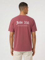 A man viewed from the back wearing a heathered dusty rose t-shirt with 'John 3:16' printed in white Gothic font on the back, paired with off-white trousers, against a light gray studio background.