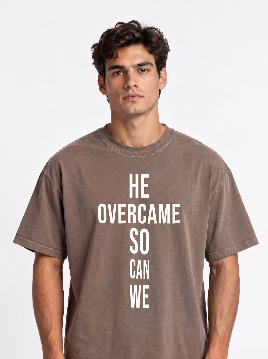 Person wearing a faded brown T-shirt with large white text reading “HE OVERCAME SO CAN WE.”