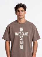 Person wearing a faded brown T-shirt with large white text reading “HE OVERCAME SO CAN WE.”