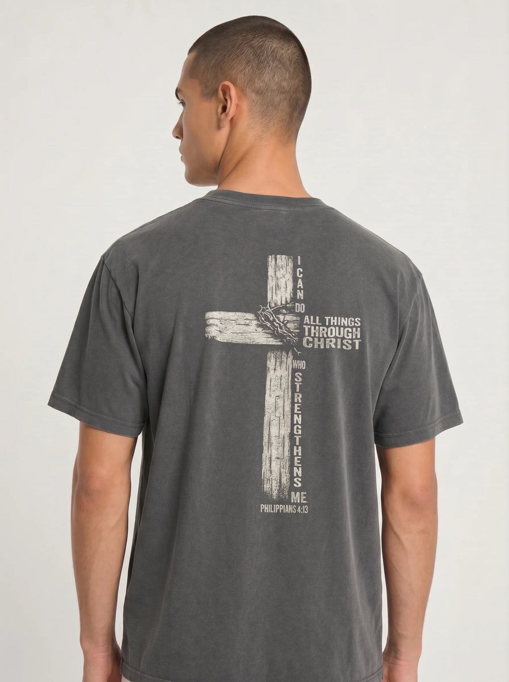 A man viewed from the back wearing a dark gray t-shirt with a distressed biege graphic of a cross with the text 'I CAN DO ALL THINGS THROUGH CHRIST WHO STRENGTHENS ME' from Philippians 4:13'.