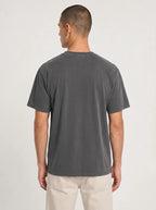 A man viewed from the back wearing a dark gray short-sleeve crewneck t-shirt and light beige pants, standing against a light gray studio background.