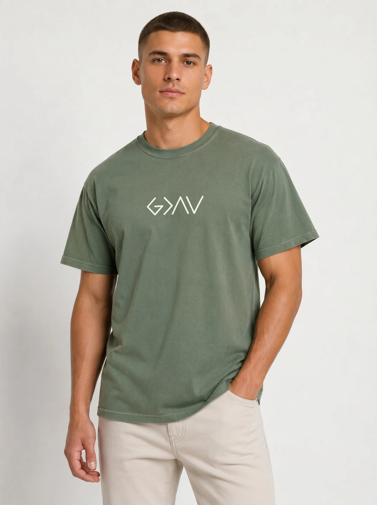 A man wearing a muted sage green crewneck t-shirt with a white stylized 'G/N/V' logo on the chest, paired with beige trousers, standing against a light gray studio background.
