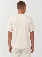 A man viewed from the back wearing a plain cream-colored short-sleeve t-shirt and matching cream pants against a light gray studio background.