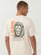 Person wearing a cream T-shirt with a lion illustration, orange “1 John 4:4” and cross, and the verse “You, dear children, are from God and have overcome them, because the one who is in you is greater than the one who is in the world.”