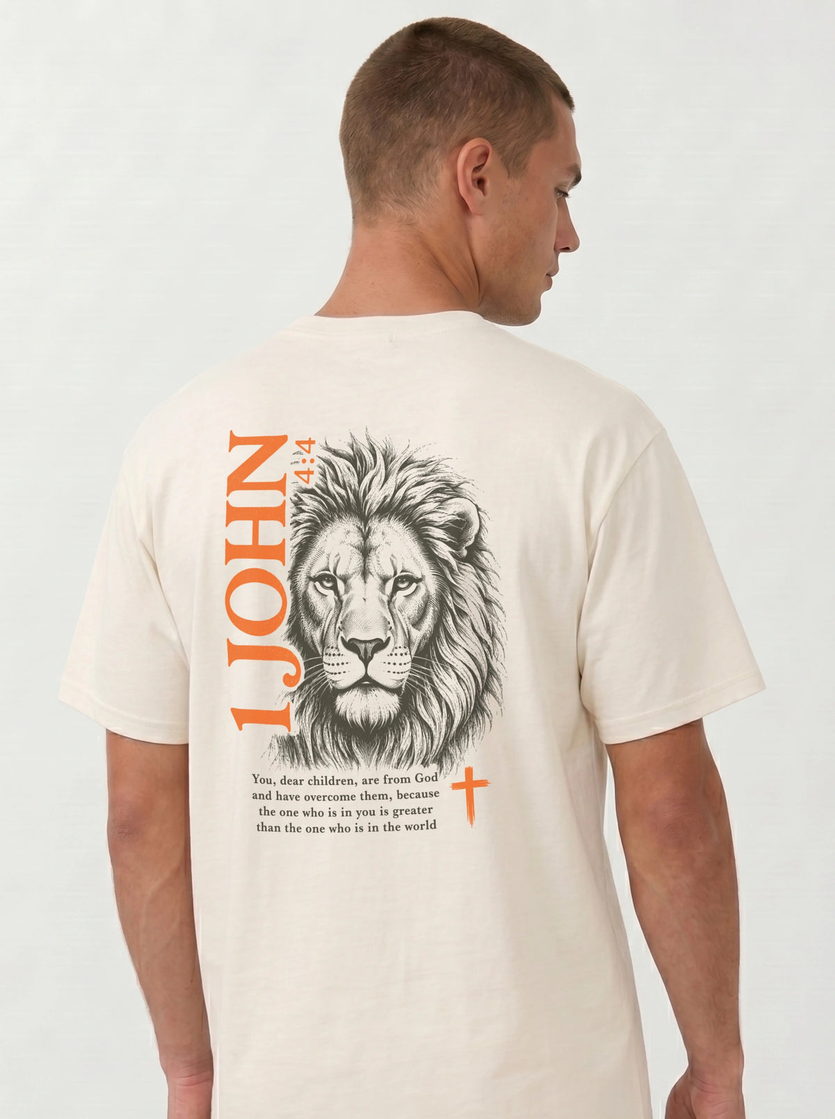 Person wearing a cream T-shirt with a lion illustration, orange “1 John 4:4” and cross, and the verse “You, dear children, are from God and have overcome them, because the one who is in you is greater than the one who is in the world.”