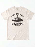 An off-white crewneck t-shirt with a black graphic print on the front. The graphic features a mountain silhouette with a cross on top, the text 'FAITH CAN MOVE MOUNTAINS', and 'MATTHEW 17:20' underneath.