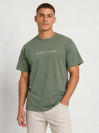 Young man modeling a sage green T-shirt featuring white “JESUS SAVES” text and a small cross on the chest, paired with light-colored pants against a plain off-white background.
