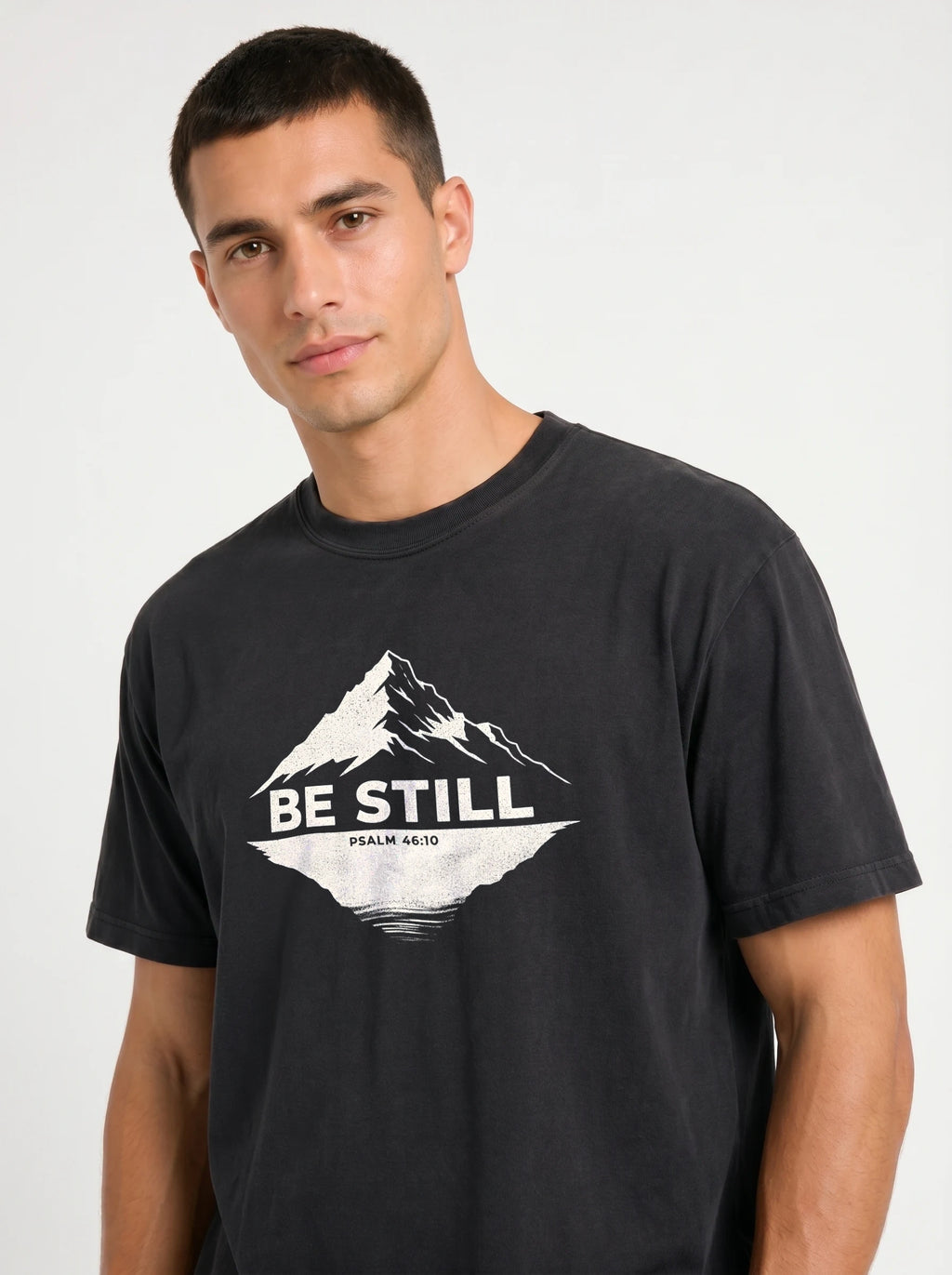 A man wearing a black graphic t-shirt with a white mountain design and the text 'BE STILL' and 'PSALM 46:10' printed on the chest, set against a plain light gray background.