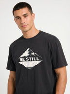 A man wearing a black graphic t-shirt with a white mountain design and the text 'BE STILL' and 'PSALM 46:10' printed on the chest, set against a plain light gray background.
