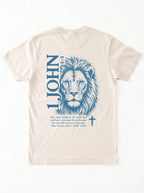 Flat lay of a cream-colored garment-dyed t-shirt, back view, laid on a white surface. It shows a large blue lion illustration with '1 JOHN 4:4' text, a Bible verse, and a small cross symbol.