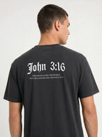 A man shown from the back wearing a black t-shirt with white gothic-style text reading 'John 3:16' and a Bible verse underneath.