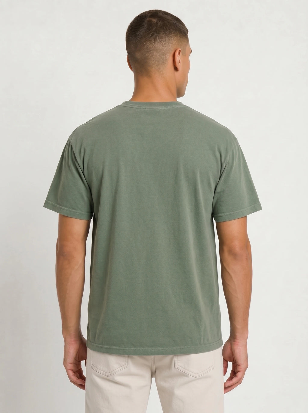 A man viewed from the back wearing a muted sage green short-sleeve t-shirt and light beige pants, standing against a light gray textured studio background.