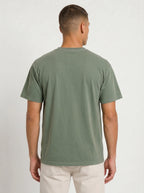 A man viewed from the back wearing a muted sage green short-sleeve t-shirt and light beige pants, standing against a light gray textured studio background.