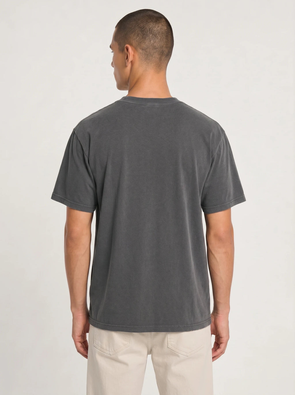 A man viewed from the back wearing a dark gray washed short-sleeve t-shirt and light beige pants, standing against a plain light gray studio background.