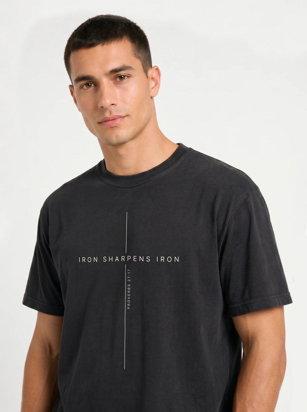 Close-up portrait of a male model looking forward, wearing a black t-shirt featuring a minimalist white cross graphic with the text "IRON SHARPENS IRON" and "PROVERBS 27:17".