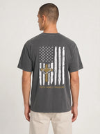 A man viewed from the back wearing a dark gray t-shirt with a distressed vertical American flag graphic on the back, featuring a gold cross and the words 'FAITH, FAMILY, FREEDOM.'.