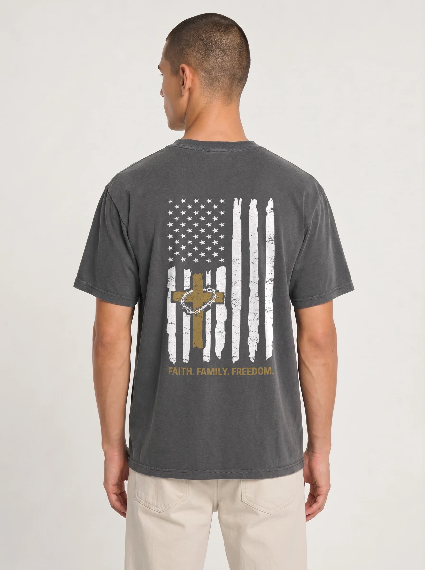 A man viewed from the back wearing a dark gray t-shirt with a distressed vertical American flag graphic on the back, featuring a gold cross and the words 'FAITH, FAMILY, FREEDOM.'.