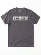 A dark gray crewneck t-shirt with 'REDEEMED' printed in distressed white block letters across the chest, flanked by two horizontal lines and a small cross below, shown flat on a white background.