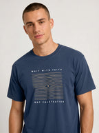 A man wearing a navy blue crewneck t-shirt withwavy line design and the text 'WAIT WITH FAITH' and 'NOT FRUSTRATION' on the front, photographed against a light gray studio background.