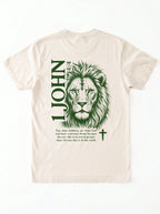 Back of a cream T-shirt with a green lion illustration, cross, “1 John 4:4,” and the verse text, “You, dear children, are from God and have overcome them, because the one who is in you is greater than the one who is in the world.”