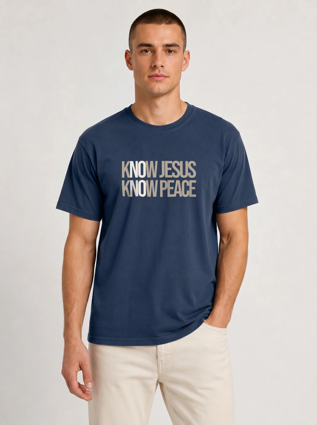 A man wearing a navy blue crewneck t-shirt with the text 'KNOW JESUS KNOW PEACE' printed in a distressed beige font on the chest, paired with light beige pants, against a light gray studio background.