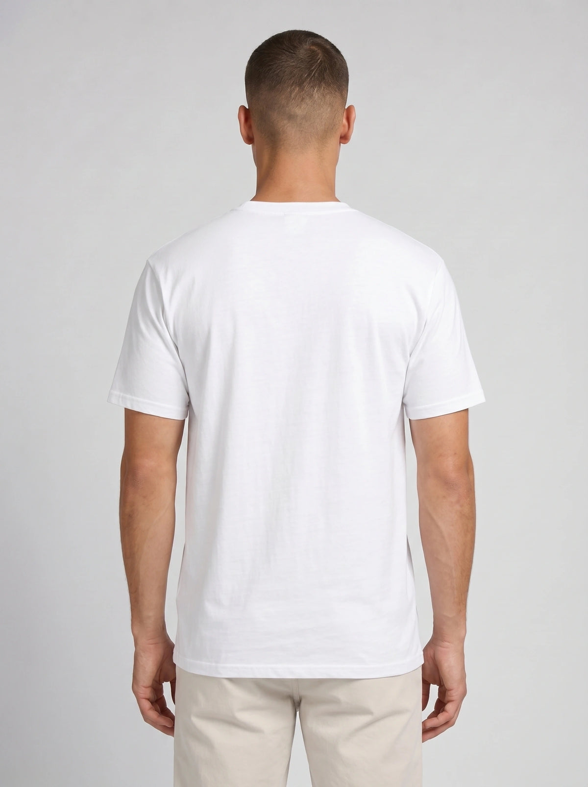 A man viewed from the back wearing a plain white short-sleeve crewneck t-shirt and light beige trousers, standing against a neutral light gray studio background.