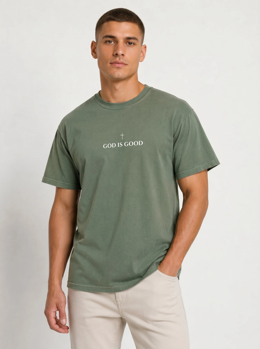 Person wearing a muted green T-shirt with a small cross above the text “God Is Good.”
