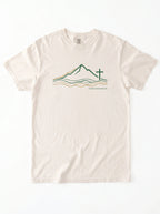 An off-white crewneck t-shirt featuring a graphic of a minimalist line art logo featuring a mountain range above flowing river lines, with a Christian cross standing on the right side. The design is drawn in green and gold on a light background, with geographic coordinates printed beneath the landscape.