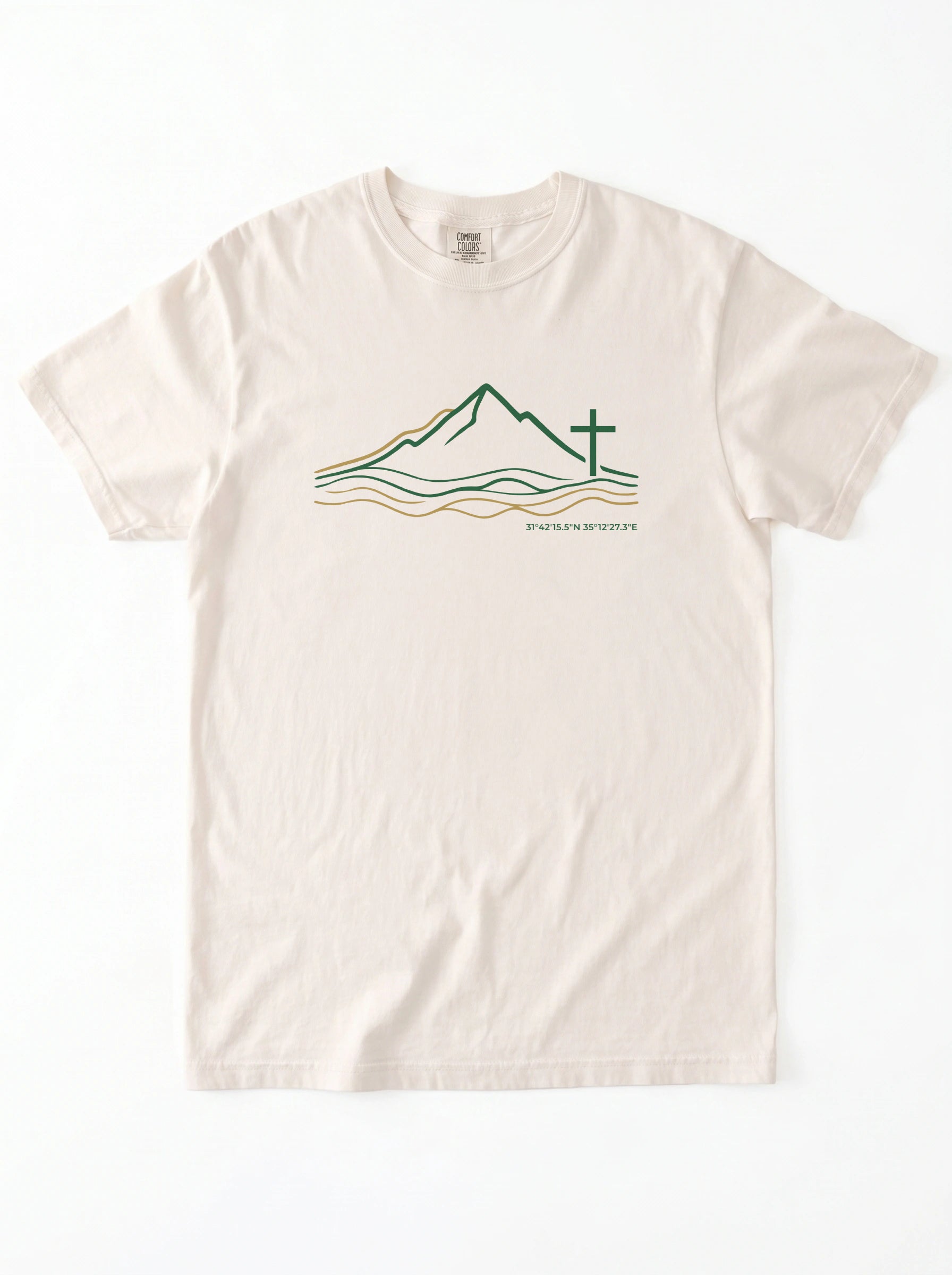 An off-white crewneck t-shirt featuring a graphic of a minimalist line art logo featuring a mountain range above flowing river lines, with a Christian cross standing on the right side. The design is drawn in green and gold on a light background, with geographic coordinates printed beneath the landscape.