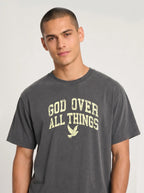 Close-up portrait of a male model looking forward, wearing a charcoal t-shirt featuring a distressed yellow graphic with the words "GOD OVER ALL THINGS" and a flying dove icon.