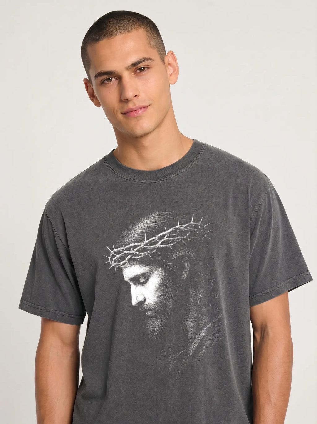 A man wearing a dark gray crewneck t-shirt with a white graphic print of Jesus' face wearing a crown of thorns, photographed against a light gray studio background.