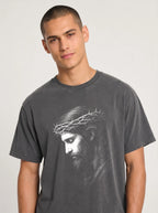 A man wearing a dark gray crewneck t-shirt with a white graphic print of Jesus' face wearing a crown of thorns, photographed against a light gray studio background.