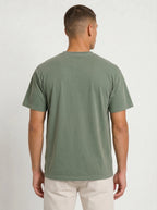 A man viewed from the back wearing a muted sage green short-sleeve t-shirt and light beige pants, standing against a plain light gray studio background.