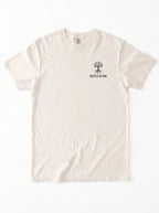 A light cream-colored short-sleeve t-shirt with a small black graphic of a tree and the text 'ROOTED IN HIM.' printed on the left chest, displayed flat against a white background.