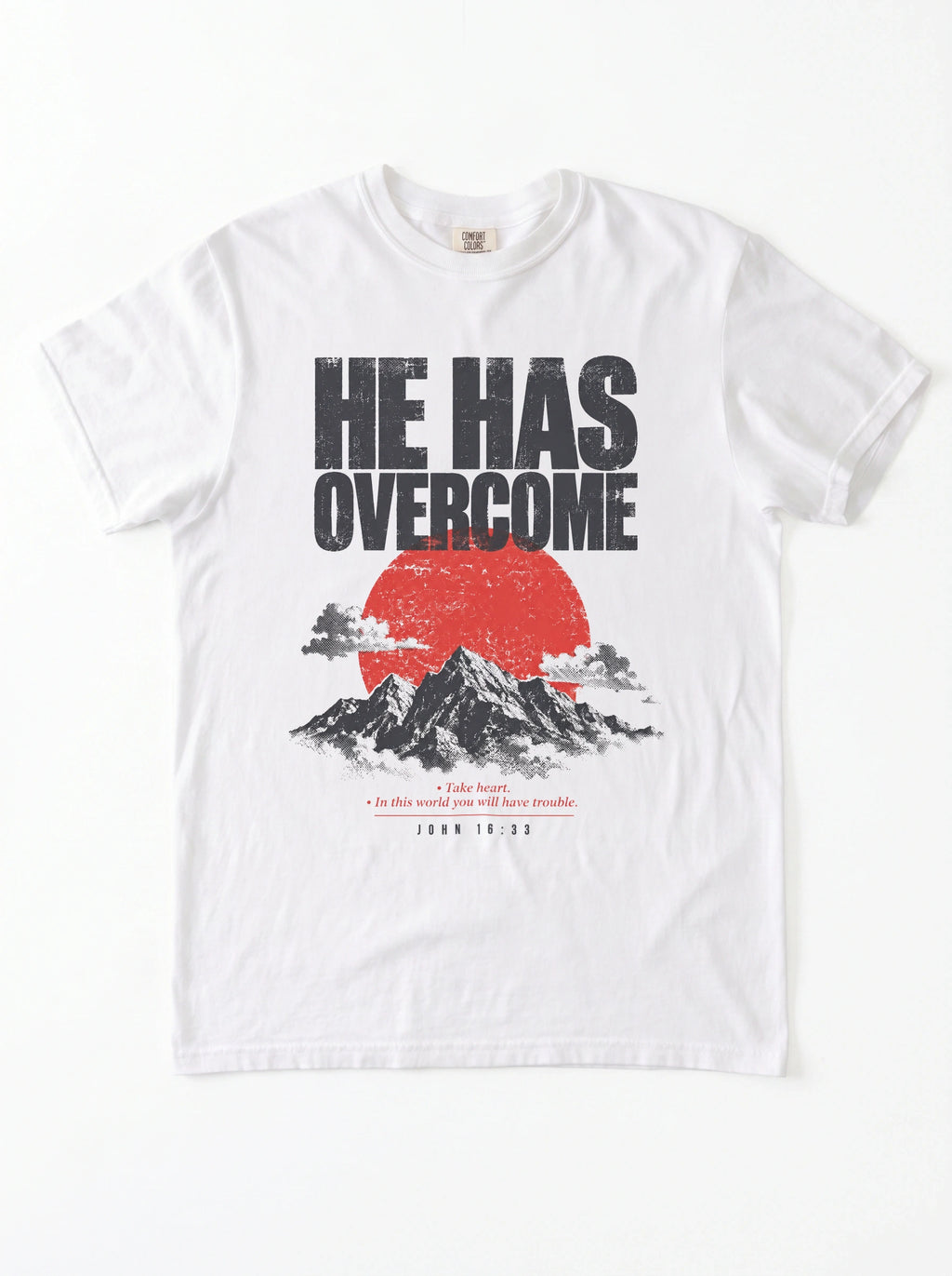 A white short-sleeve crewneck t-shirt featuring a graphic print on the front with the text 'HE HAS OVERCOME' above a red sun and black mountain illustration, with a Bible verse printed below.
