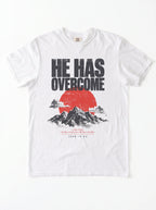 A white short-sleeve crewneck t-shirt featuring a graphic print on the front with the text 'HE HAS OVERCOME' above a red sun and black mountain illustration, with a Bible verse printed below.