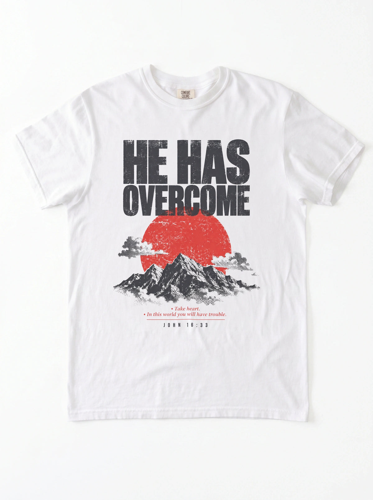 A white short-sleeve crewneck t-shirt featuring a graphic print on the front with the text 'HE HAS OVERCOME' above a red sun and black mountain illustration, with a Bible verse printed below.