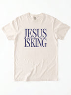 Cream T-shirt laid flat with distressed navy text reading “Jesus Is King.”