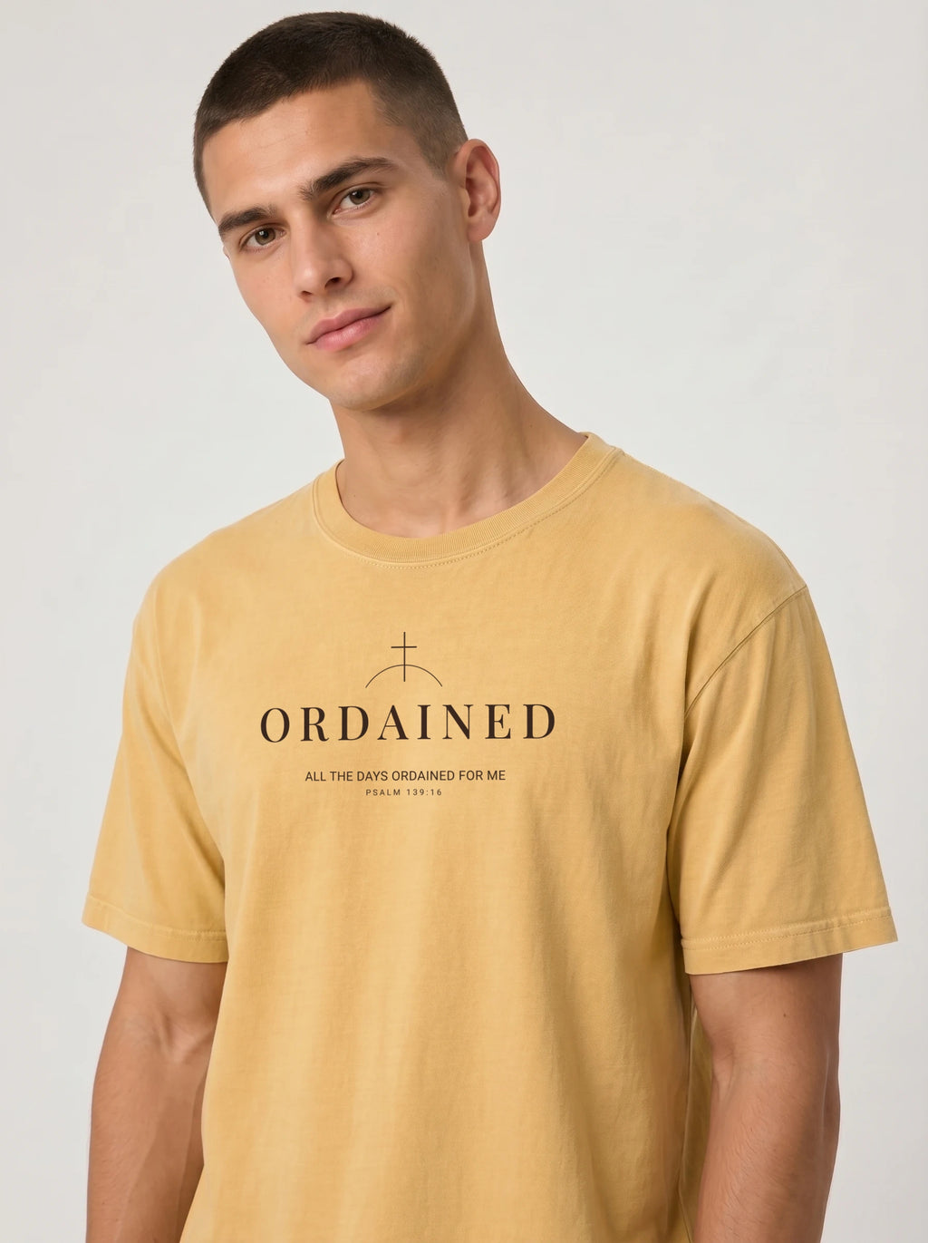 A man wearing a mustard yellow t-shirt with a black printed graphic on the chest that reads 'ORDAINED' with a cross symbol and scripture text, set against a plain light gray background.
