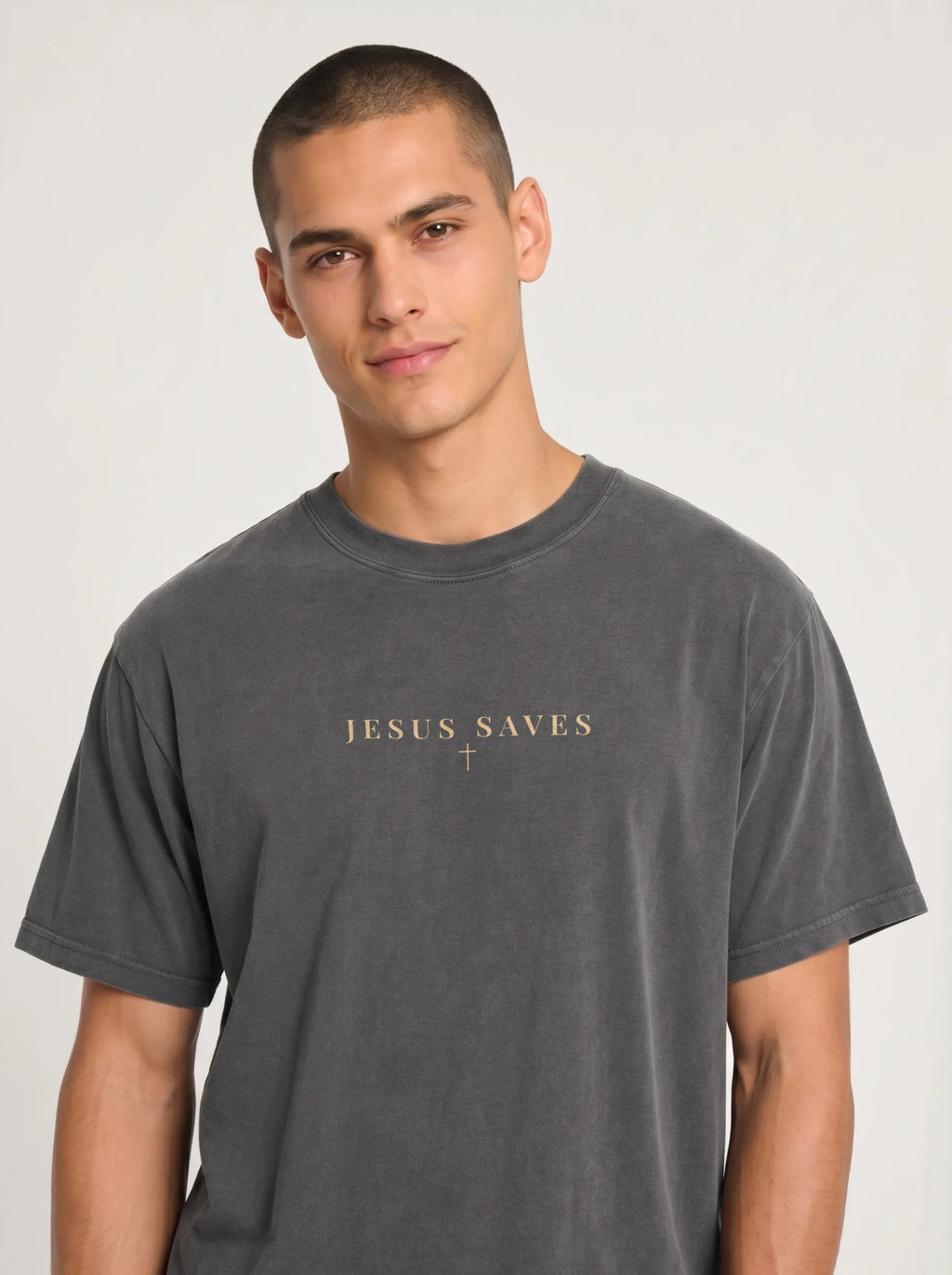 Young man with a buzz cut wearing an oversized charcoal-gray T-shirt with the words “JESUS SAVES” and a small cross printed on the chest, standing against a plain light background.