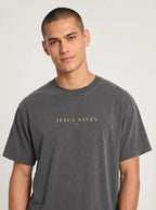 Young man with a buzz cut wearing an oversized charcoal-gray T-shirt with the words “JESUS SAVES” and a small cross printed on the chest, standing against a plain light background.