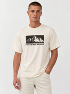 A man wearing a cream-colored crewneck t-shirt with a black graphic of mountains and the text 'FAITH CAN MOVE MOUNTAINS' on the chest, paired with matching cream pants, against a light gray studio background.
