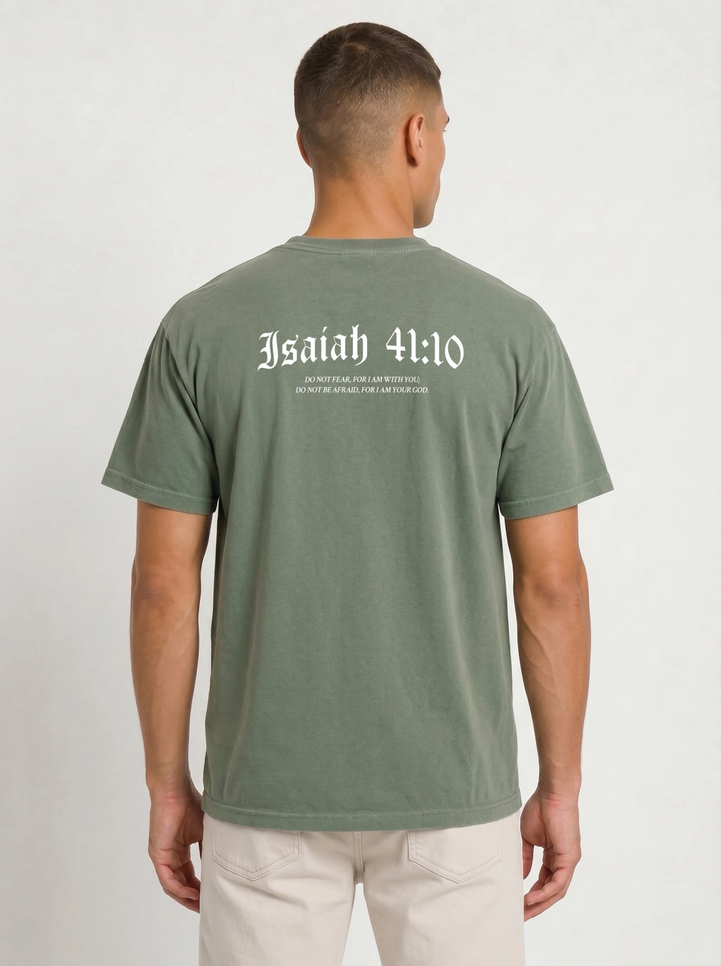 A man viewed from the back wearing an olive green t-shirt with 'Isaiah 41:10' printed in white Gothic lettering across the upper back, paired with off-white pants, against a plain light gray background.