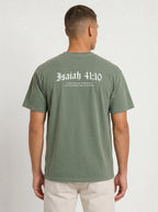 A man viewed from the back wearing an olive green t-shirt with 'Isaiah 41:10' printed in white Gothic lettering across the upper back, paired with off-white pants, against a plain light gray background.