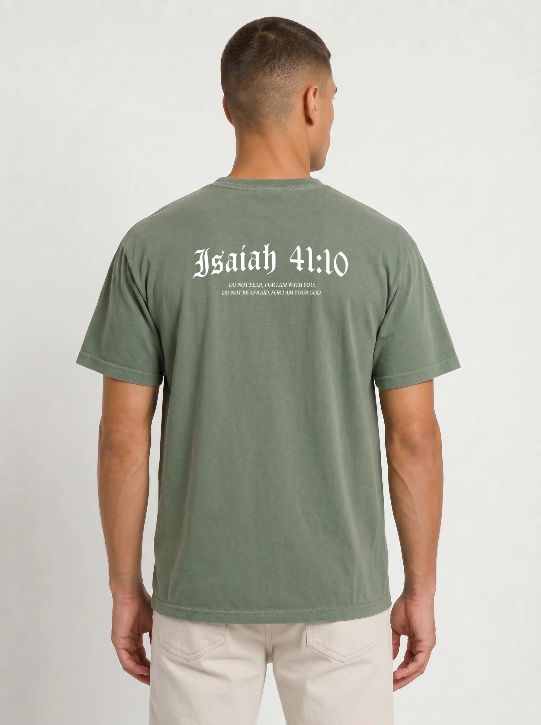 A man viewed from the back wearing an olive green t-shirt with 'Isaiah 41:10' printed in white Gothic lettering across the upper back, paired with off-white pants, against a plain light gray background.