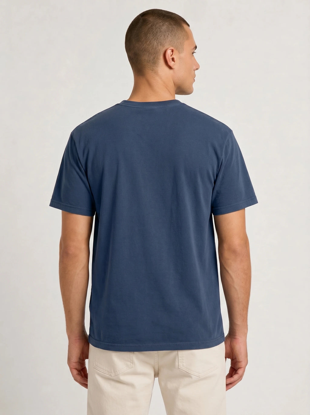 A man viewed from the back wearing a solid navy blue short-sleeve crewneck t-shirt and light beige trousers, set against a plain light gray studio background.