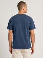 A man viewed from the back wearing a solid navy blue short-sleeve crewneck t-shirt and light beige trousers, set against a plain light gray studio background.