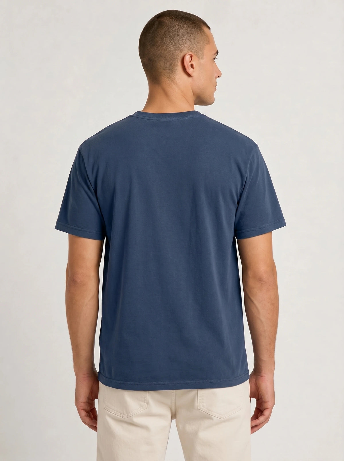 A man viewed from the back wearing a solid navy blue short-sleeve crewneck t-shirt and light beige trousers, set against a plain light gray studio background.